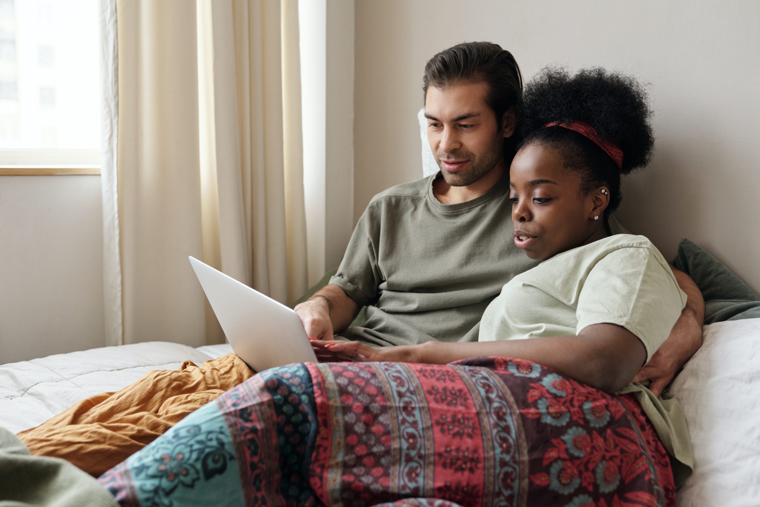 Multiracial couple sitting in bed together while watching something on a laptop